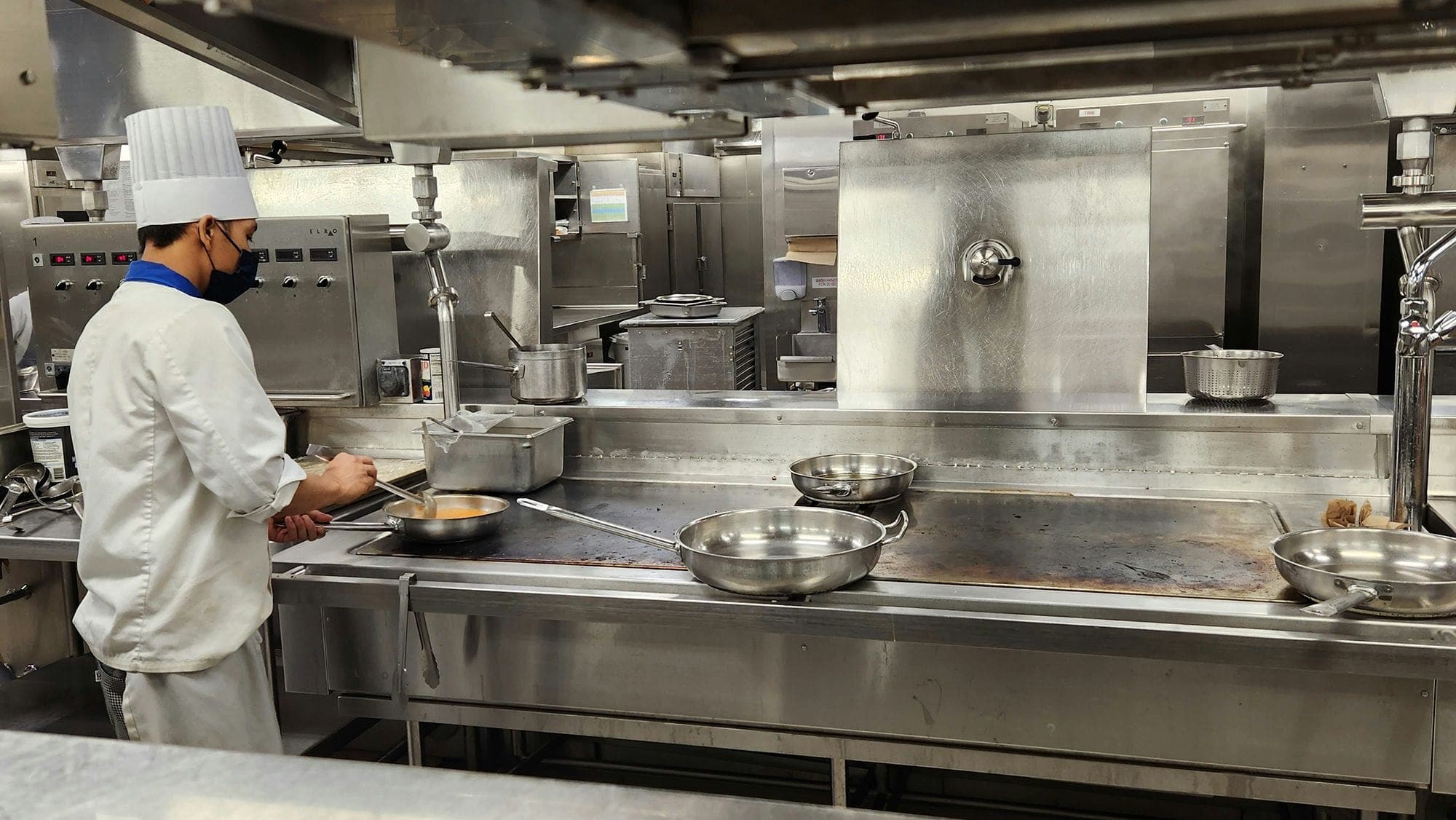 Photograph of a chef in a restaurant kitchen, cooking something in a pan over a heated stovetop.