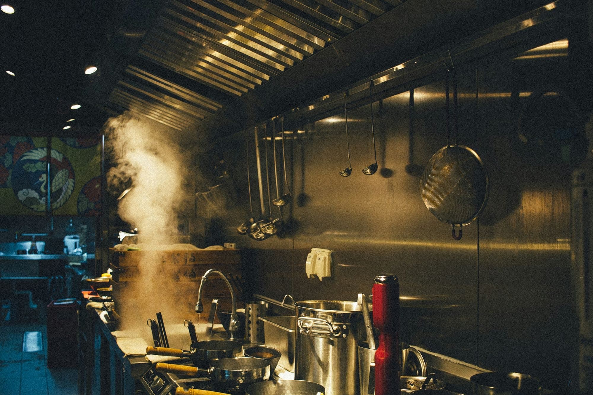 Photograph of a dimly lit restaurant kitchen; hanging on the wall are a series of ladles and strainers and on the counter are a series of pots and pans. The back half of the counter is obscured by steam.